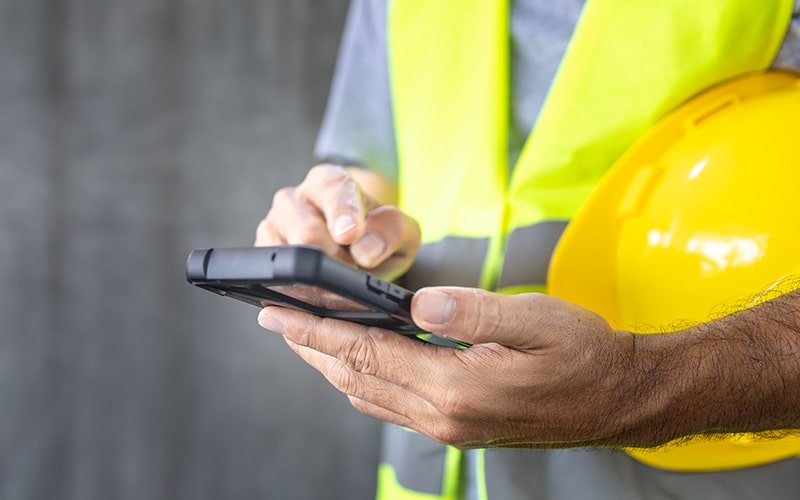 un responsabile del servizio di prevenzione e prevenzione che indossa un gilet ad alta visibilità e tiene in mano un elmetto di sicurezza giallo mentre usa uno smartphone