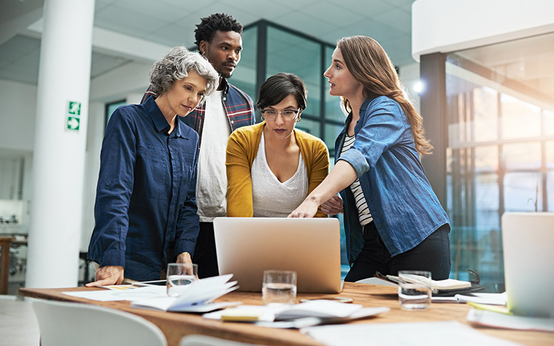 Quattro colleghi al lavoro intorno a un laptop, che discutono le differenze tra lavoro supplementare e lavoro straordinario.